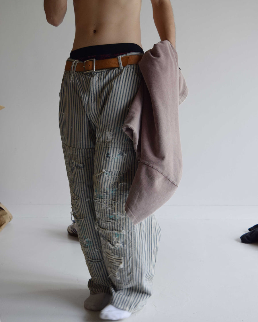 過去の定番ダブルニーペインターパンツをリファインし、腰回りにゆとりを持たせたワイドストレートシルエットに仕上げたヒッコリーパンツ。
岡山県井原市製の9ozヒッコリーストライプ生地を使用し、ハリのある風合いが特徴。
全体にエイジングとハードダメージを施し、手作業のステッチ補修やランダムなペンキでリアルな表情を加えています。