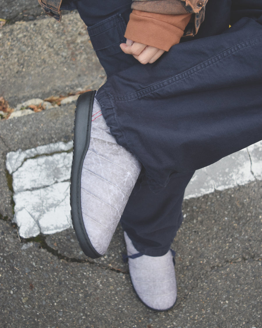 リラックスサンダル〈SUBU〉とのコラボレーションによって生まれた、特別仕様のフットギア。
アッパーにはANACHRONORMが独自の加工を施したナイロンオックスを採用。
染料の濃度や色味を幾度も検証し、洗いをかけることで偶発的なムラや陰影が生まれ、ひとつひとつランダムな表情を持つ一足が完成しました。
ふんわりと包み込む起毛ライニングと4層構造のインソールが、ダウンのようなぬくもりと心地よい弾力を生み出します。
SUBUの機能性にANACHRONORMのクラフト感が融合した、冬の定番を新たに更新する一足です。
カラーはホワイト。