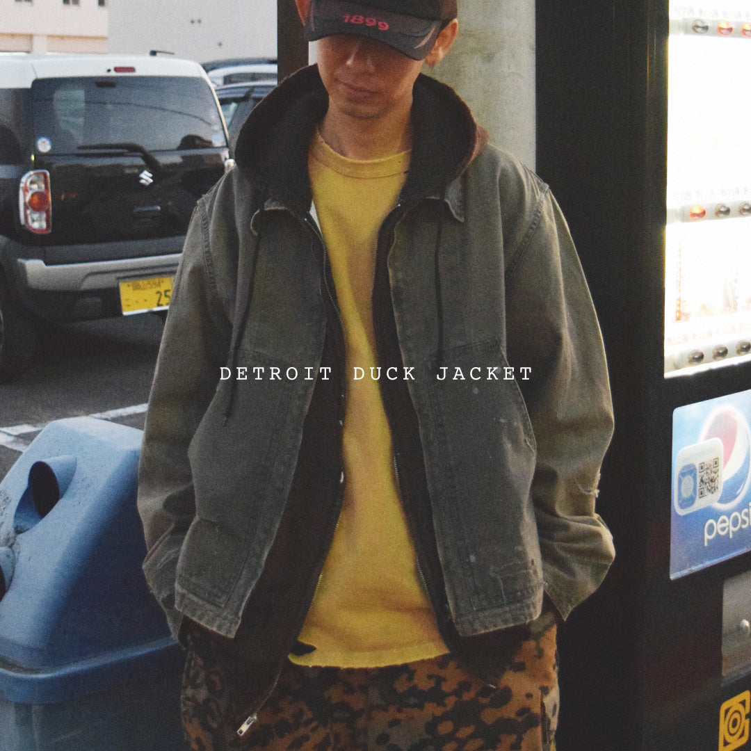 【 商品名：リメイクデトロイトダックジャケット 】本製品は、ゆとりのある身幅とややクロップド丈に設計されたブラックカラーのジャケットで、インナーアイテムとのレイヤードを楽しめるバランスの良いシルエットが特徴です。生地には頒布の中でも最も薄手の11号帆布生地を採用しており、強度・耐久性に優れ、着用を重ねるごとにアタリなどの経年変化を味わえる素材です。さらに、水や薬品の使用量を抑えながら生地の凹凸部分のアタリを強調し、同時に自然な脱色感を生み出す特殊エイジング加工を施し、加えてサンフェードによるダメージ加工を加えることで、より奥行きのある表情に仕上げています。
