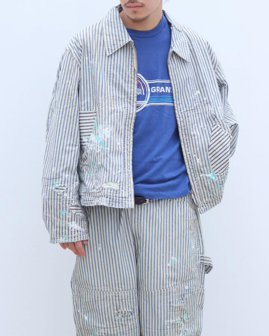 ゆとりある身幅とややクロップド丈でレイヤードを楽しめるデザインにしたヒッコリージャケット。
岡山県製9ozヒッコリーストライプ生地を使用し、ハリのあるしっかりとした風合いが特徴。
全体にエイジングとハードダメージを施し、手作業のステッチ補修とランダムなペンキでリアルな表情に仕上げています。