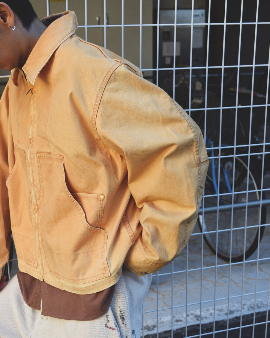 シルエットはゆとりのある身幅とややクロップド丈で設計しており、イエローカラーの鮮やかなトーンがレイヤードスタイルをより引き立てます。インナーアイテムとの重ね着がしやすいバランスに仕上げています。
生地には頒布の中で最も薄手の11号帆布を採用しており、強度・耐久性に優れるだけでなく、着用を重ねるごとにアタリなどの経年変化を楽しめる素材です。また、特殊エイジング加工を施すことで、水や薬品の使用量を抑えながら生地の凹凸が強調され、イエローの色味に自然なフェード感を加えています。