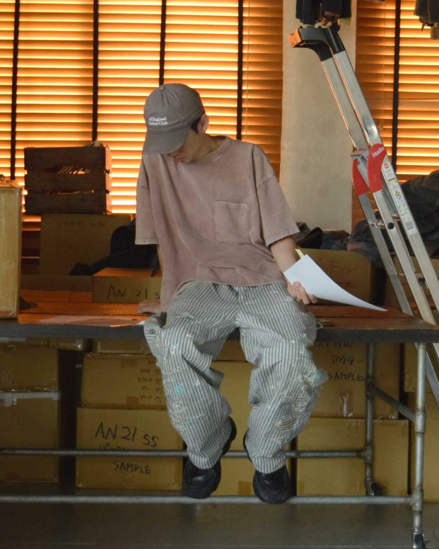 過去の定番ダブルニーペインターパンツをリファインし、腰回りにゆとりを持たせたワイドストレートシルエットに仕上げたヒッコリーパンツ。
岡山県井原市製の9ozヒッコリーストライプ生地を使用し、ハリのある風合いが特徴。
全体にエイジングとハードダメージを施し、手作業のステッチ補修やランダムなペンキでリアルな表情を加えています。