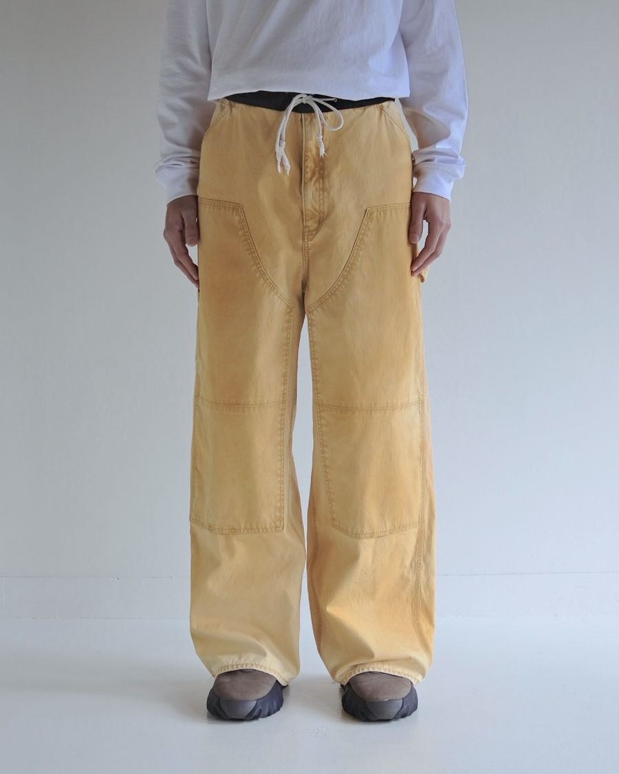 【 商品名：ダブルニーダックペインターパンツ 】

本製品は、レングスをやや長めに設定したイエローカラーのペインターパンツです。ウエスト部分にはナイロンオックス生地で切り替えを施し、ワーク感を抑えたデザインに仕上げています。生地には頒布の中でも最も薄手の11号帆布生地を使用しており、強度・耐久性に優れるだけでなく、着用を重ねるごとにアタリなどの経年変化を楽しめる素材です。また、水や薬品の使用量を抑えつつ生地の凹凸部分のアタリを強調し、同時に自然な脱色感を生み出す特殊エイジング加工を施しています。