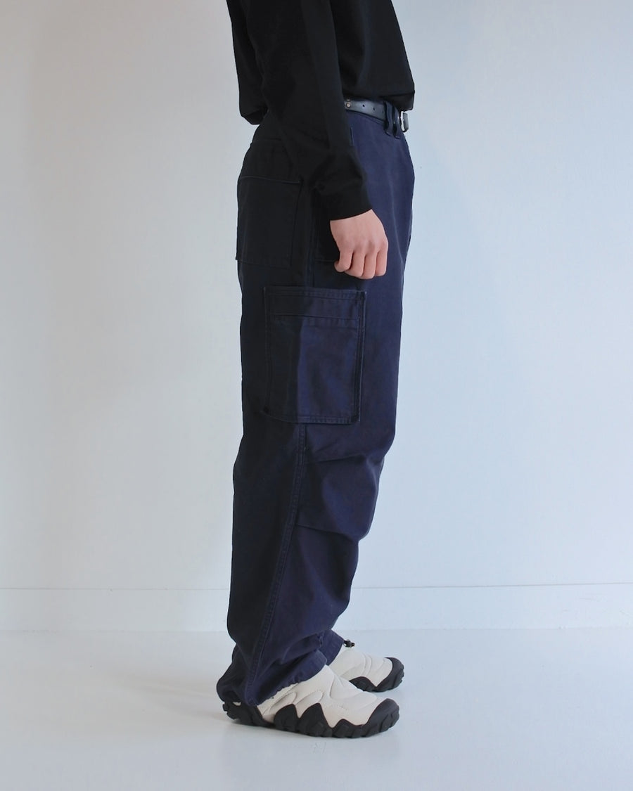 フラップポケットや膝のタック、裾のドローコードなど、ミリタリーディテールを随所に取り入れた1本。通常は内側に配置されるポケットをあえて外側に設けたカーゴポケットが印象的です。ムラ糸で織り上げたバックサテン生地は、独特のハリとコシを持ち、深みのあるネイビーカラーが魅力です。