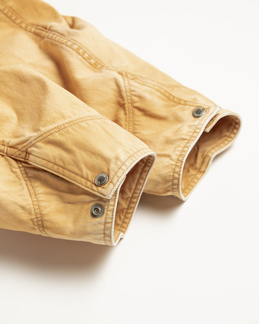 シルエットはゆとりのある身幅とややクロップド丈で設計しており、イエローカラーの鮮やかなトーンがレイヤードスタイルをより引き立てます。インナーアイテムとの重ね着がしやすいバランスに仕上げています。
生地には頒布の中で最も薄手の11号帆布を採用しており、強度・耐久性に優れるだけでなく、着用を重ねるごとにアタリなどの経年変化を楽しめる素材です。また、特殊エイジング加工を施すことで、水や薬品の使用量を抑えながら生地の凹凸が強調され、イエローの色味に自然なフェード感を加えています。