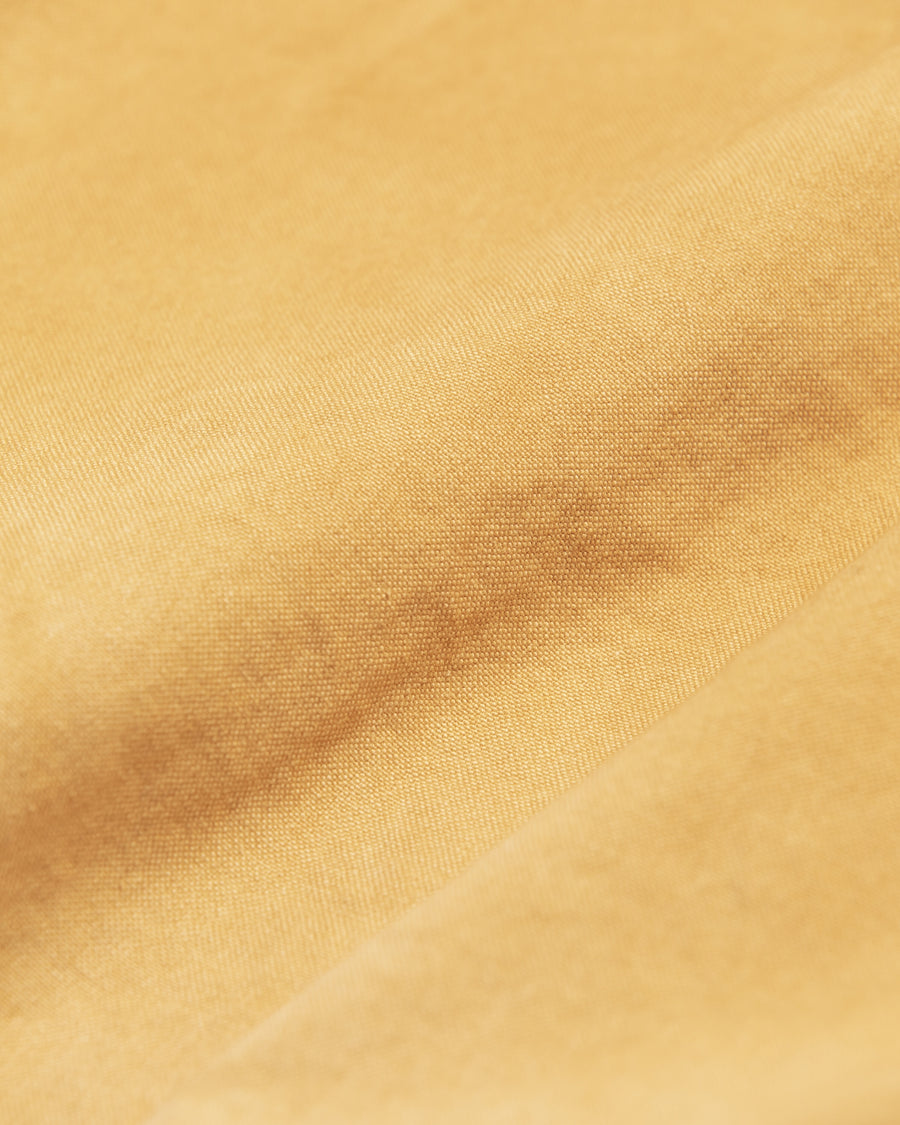 シルエットはゆとりのある身幅とややクロップド丈で設計しており、イエローカラーの鮮やかなトーンがレイヤードスタイルをより引き立てます。インナーアイテムとの重ね着がしやすいバランスに仕上げています。
生地には頒布の中で最も薄手の11号帆布を採用しており、強度・耐久性に優れるだけでなく、着用を重ねるごとにアタリなどの経年変化を楽しめる素材です。また、特殊エイジング加工を施すことで、水や薬品の使用量を抑えながら生地の凹凸が強調され、イエローの色味に自然なフェード感を加えています。