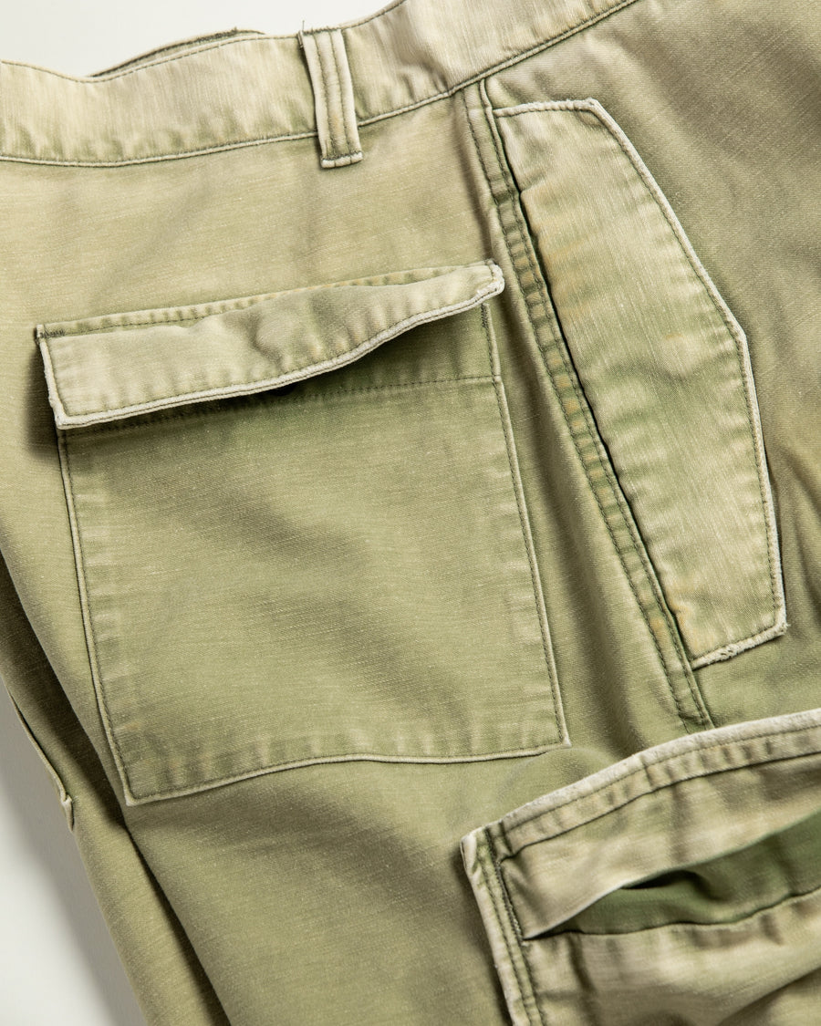 フラップポケットや膝のタック、裾のドローコードなど、ミリタリーのディテールをふんだんに取り入れた一本。内側に配置されることの多いポケットをあえて外側に設けたカーゴポケットが特徴です。ムラ糸で織り上げたバックサテン生地は、ハリとコシのある質感が魅力。さらに、水や薬品の使用を抑えながら生地の凹凸を際立たせる特殊エイジング加工を施し、深みのあるオリーブカラーに仕上げています。