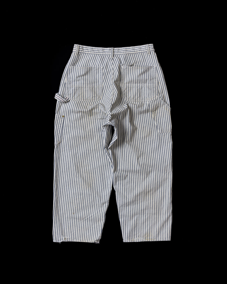 過去の定番ダブルニーペインターパンツをリファインし、腰回りにゆとりを持たせたワイドストレートシルエットに仕上げたヒッコリーパンツ。
岡山県井原市製の9ozヒッコリーストライプ生地を使用し、ハリのある風合いが特徴。
全体にエイジングとハードダメージを施し、手作業のステッチ補修やランダムなペンキでリアルな表情を加えています。