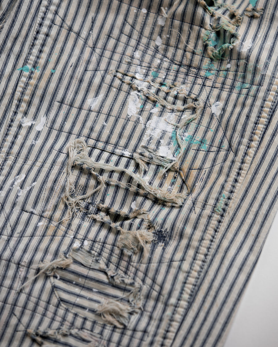 過去の定番ダブルニーペインターパンツをリファインし、腰回りにゆとりを持たせたワイドストレートシルエットに仕上げたヒッコリーパンツ。
岡山県井原市製の9ozヒッコリーストライプ生地を使用し、ハリのある風合いが特徴。
全体にエイジングとハードダメージを施し、手作業のステッチ補修やランダムなペンキでリアルな表情を加えています。