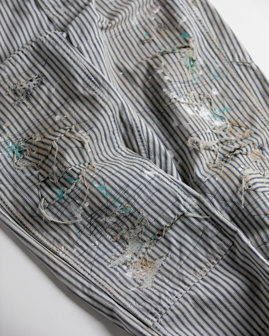 過去の定番ダブルニーペインターパンツをリファインし、腰回りにゆとりを持たせたワイドストレートシルエットに仕上げたヒッコリーパンツ。
岡山県井原市製の9ozヒッコリーストライプ生地を使用し、ハリのある風合いが特徴。
全体にエイジングとハードダメージを施し、手作業のステッチ補修やランダムなペンキでリアルな表情を加えています。