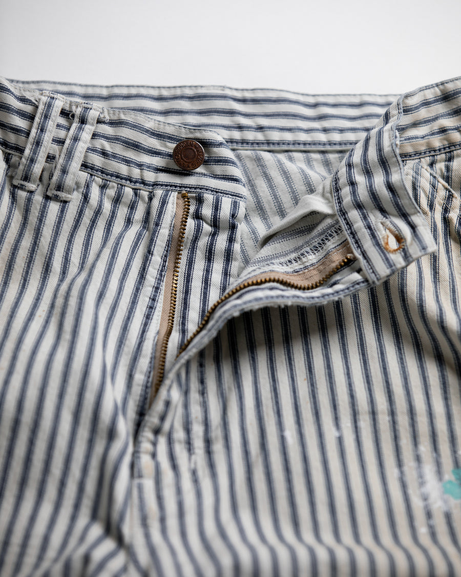 過去の定番ダブルニーペインターパンツをリファインし、腰回りにゆとりを持たせたワイドストレートシルエットに仕上げたヒッコリーパンツ。
岡山県井原市製の9ozヒッコリーストライプ生地を使用し、ハリのある風合いが特徴。
全体にエイジングとハードダメージを施し、手作業のステッチ補修やランダムなペンキでリアルな表情を加えています。