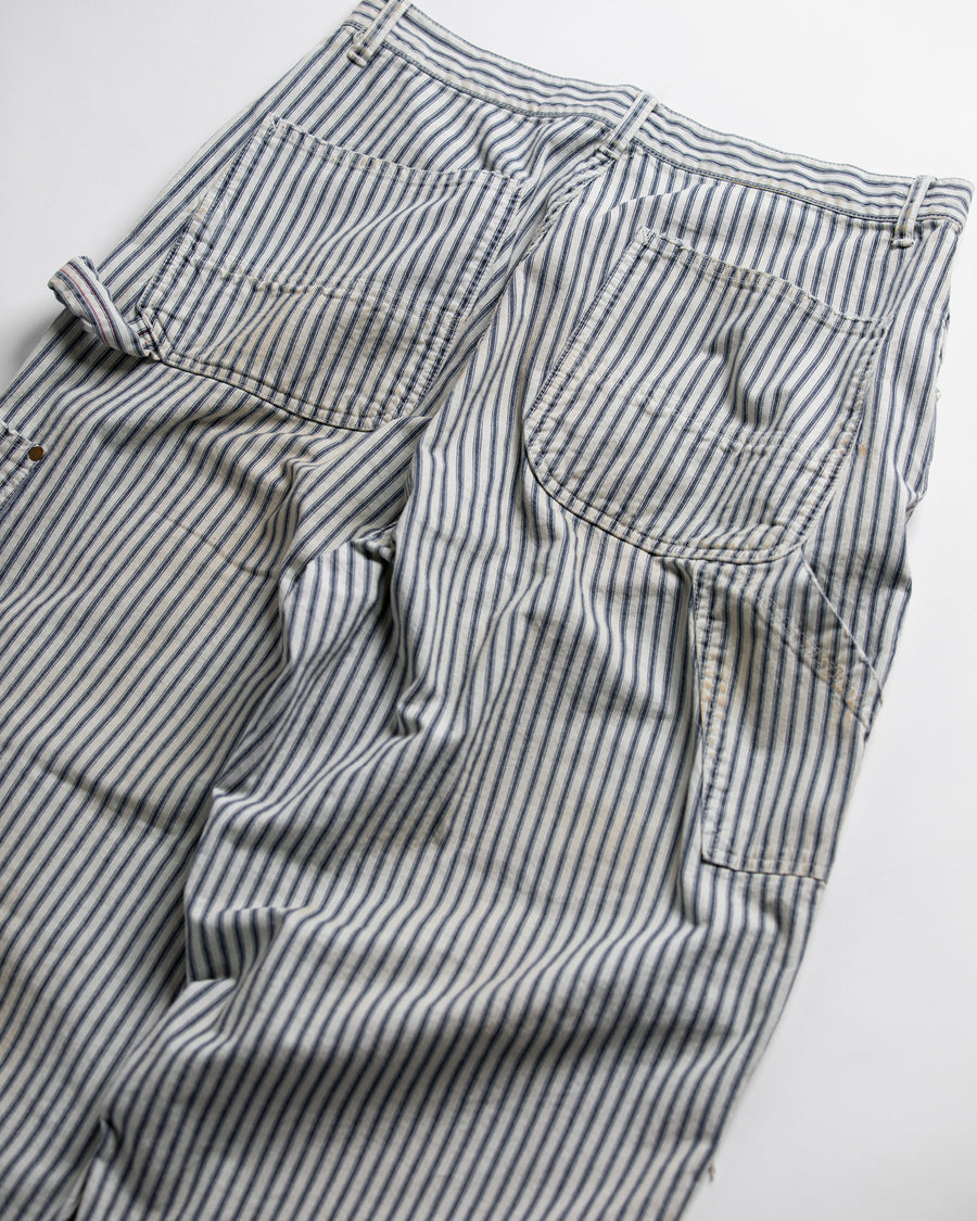 過去の定番ダブルニーペインターパンツをリファインし、腰回りにゆとりを持たせたワイドストレートシルエットに仕上げたヒッコリーパンツ。
岡山県井原市製の9ozヒッコリーストライプ生地を使用し、ハリのある風合いが特徴。
全体にエイジングとハードダメージを施し、手作業のステッチ補修やランダムなペンキでリアルな表情を加えています。