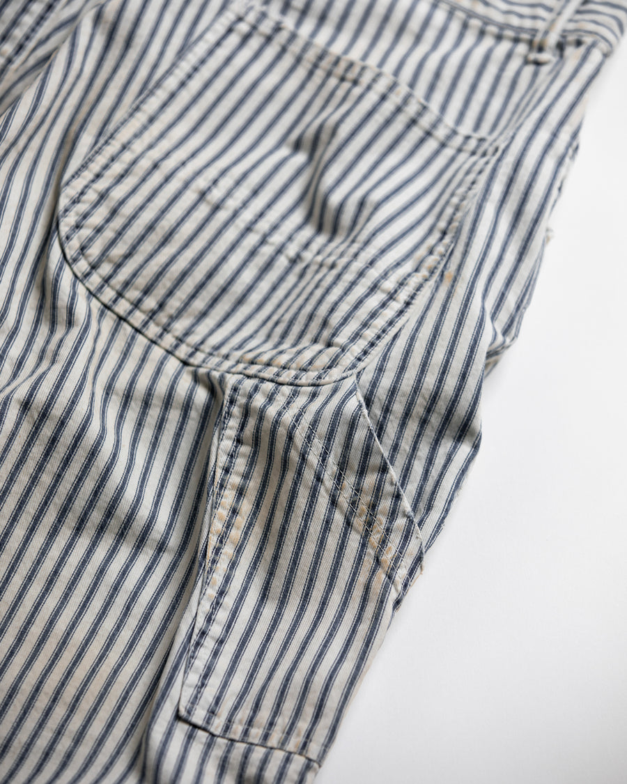 過去の定番ダブルニーペインターパンツをリファインし、腰回りにゆとりを持たせたワイドストレートシルエットに仕上げたヒッコリーパンツ。
岡山県井原市製の9ozヒッコリーストライプ生地を使用し、ハリのある風合いが特徴。
全体にエイジングとハードダメージを施し、手作業のステッチ補修やランダムなペンキでリアルな表情を加えています。