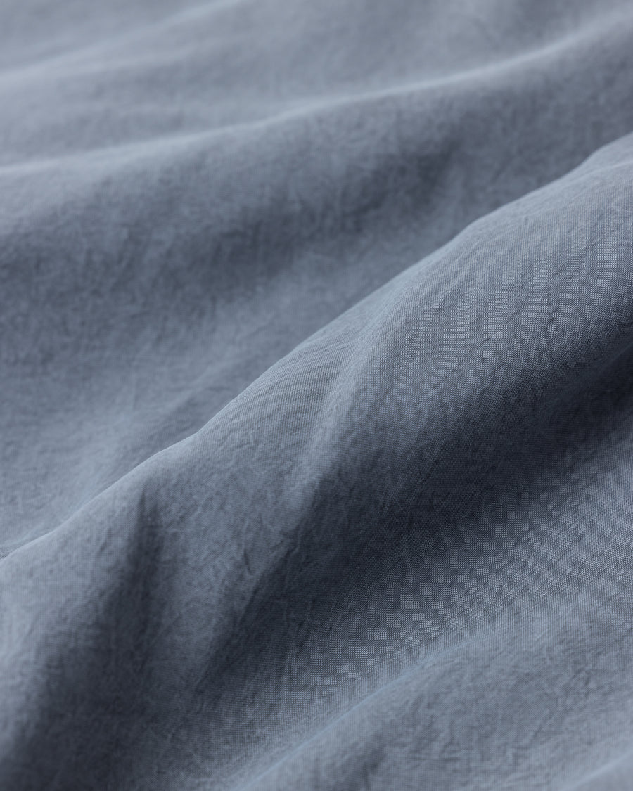 ポリエステル100％でありながら、綿のような質感と軽量性を備えた高機能な一着。タスラン加工糸を使用したポリエステルオックスを採用し、シワになりにくく速乾性に優れたイージーケア性を実現しました。本来染色が困難な素材に特殊顔料染めを施すことで、化繊特有の光沢を抑えた、ナチュラルで奥行きのある絶妙なフェード感を表現しています。

A high-performance piece crafted from polyester oxford, offering a cotton-like touch with lightweight, quick-dry, and wrinkle-resistant properties. This GRAY BLUE item features a special pigment dye—a rare process for polyester—achieving a sophisticated, faded texture and a matte finish that defies its synthetic origins.