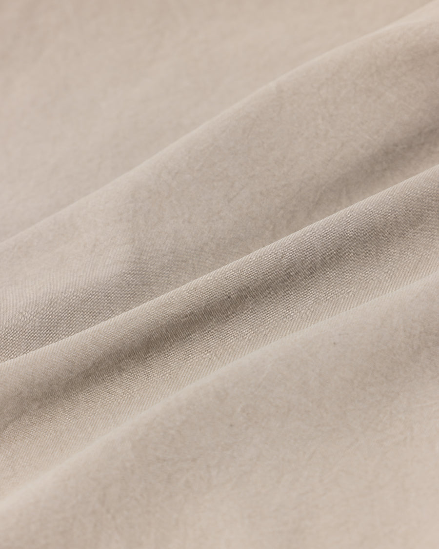 ポリエステル100％ながら、綿のような質感と軽量性を備えた高機能素材を採用。タスラン加工糸を使用したポリエステルオックスは、シワになりにくく速乾性にも優れています。本来染色が困難な素材に特殊顔料染めを施すことで、化繊特有の光沢を抑え、ナチュラルで奥行きのある絶妙なフェード感と落ち着いた表情を実現しました。

A high-performance piece crafted from polyester oxford, offering a cotton-like touch with quick-dry and wrinkle-resistant benefits. This GRAY BEIGE item features a special pigment dye—a rare process for polyester—achieving a sophisticated, faded texture and a matte finish that perfectly balances technical utility with a natural, lived-in aesthetic.