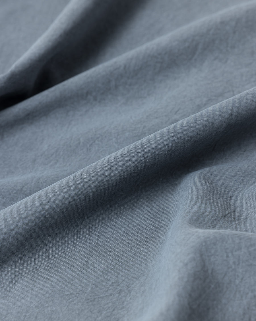 ポリエステル100％ながら、綿のような質感と軽量性を備えた高機能素材を採用。タスラン加工糸を使用したポリエステルオックスは、シワになりにくく速乾性にも優れています。本来染色が困難な素材に特殊顔料染めを施すことで、化繊特有の光沢を抑え、ナチュラルで奥行きのある絶妙なフェード感と落ち着いた表情を実現しました。

A high-performance piece crafted from polyester oxford, offering a cotton-like touch with quick-dry and wrinkle-resistant benefits. This GRAY BLUE item features a special pigment dye—a rare process for polyester—achieving a sophisticated, faded texture and a matte finish that perfectly balances technical utility with a natural, lived-in aesthetic.