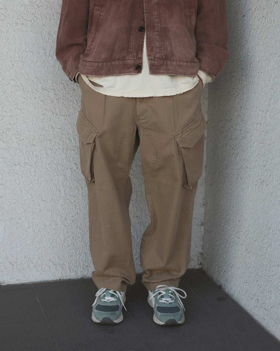 ミリタリートラウザーズをベースに、シルエットを現代的にリファイン。印象的な斜めのカーゴポケットが、立体感と実用性を両立させています。裾にかけて緩やかに絞られたテーパードラインにより、無骨ながらも洗練された印象に。素材には肌馴染みの良い10sヘリンボンを採用し、ストーンバイオウォッシュで柔らかな風合いとこなれ感を引き出しました。

Military trousers refined with a modern tapered silhouette. The distinctive slanted cargo pockets offer both practical utility and a structured look. Crafted from soft, classic 10s herringbone, this KHAKI pair has been treated with a stone bio-wash, providing a comfortable, worn-in texture and a sophisticated take on rugged style.
