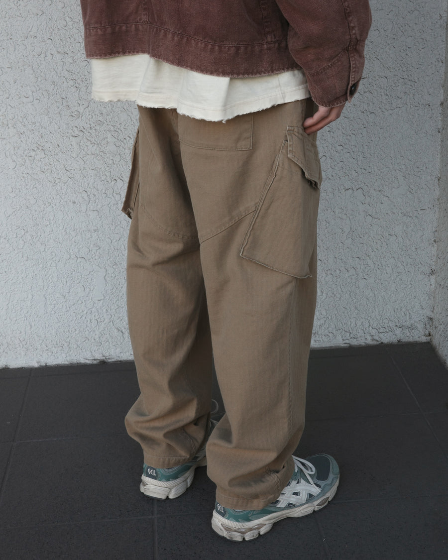 ミリタリートラウザーズをベースに、シルエットを現代的にリファイン。印象的な斜めのカーゴポケットが、立体感と実用性を両立させています。裾にかけて緩やかに絞られたテーパードラインにより、無骨ながらも洗練された印象に。素材には肌馴染みの良い10sヘリンボンを採用し、ストーンバイオウォッシュで柔らかな風合いとこなれ感を引き出しました。

Military trousers refined with a modern tapered silhouette. The distinctive slanted cargo pockets offer both practical utility and a structured look. Crafted from soft, classic 10s herringbone, this KHAKI pair has been treated with a stone bio-wash, providing a comfortable, worn-in texture and a sophisticated take on rugged style.