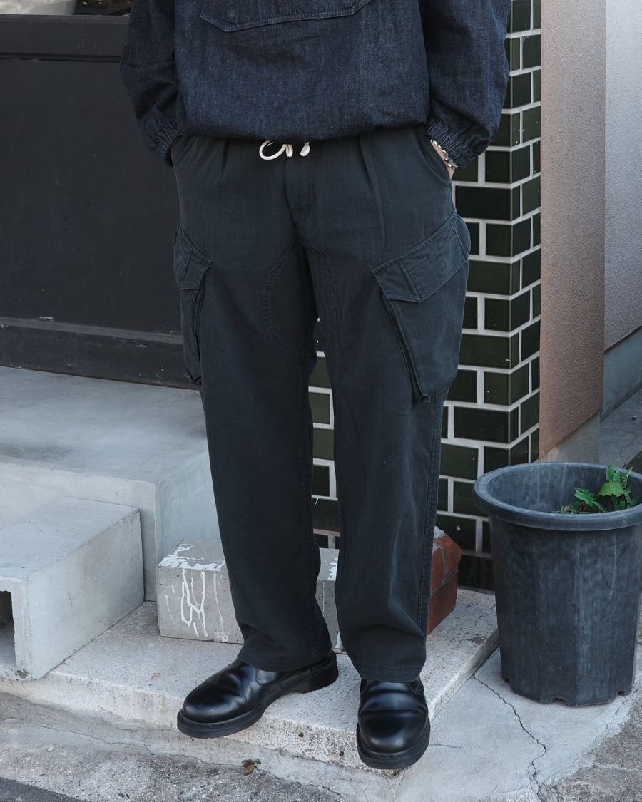 ミリタリートラウザーズをベースに、シルエットを現代的にリファイン。印象的な斜めのカーゴポケットが、立体感と実用性を両立させています。裾にかけて緩やかに絞られたテーパードラインにより、無骨ながらも洗練された印象に。素材には肌馴染みの良い10sヘリンボンを採用し、ストーンバイオウォッシュで柔らかな風合いとこなれ感を引き出しました。

Military trousers refined with a modern tapered silhouette. The distinctive slanted cargo pockets offer both practical utility and a structured look. Crafted from soft, classic 10s herringbone, this BLACK pair has been treated with a stone bio-wash, providing a comfortable, worn-in texture and a sophisticated take on rugged style.