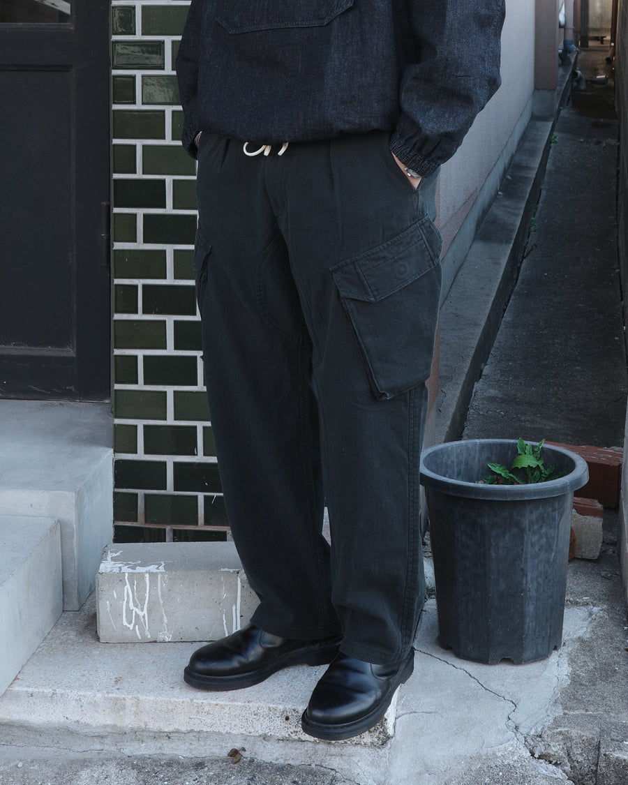 ミリタリートラウザーズをベースに、シルエットを現代的にリファイン。印象的な斜めのカーゴポケットが、立体感と実用性を両立させています。裾にかけて緩やかに絞られたテーパードラインにより、無骨ながらも洗練された印象に。素材には肌馴染みの良い10sヘリンボンを採用し、ストーンバイオウォッシュで柔らかな風合いとこなれ感を引き出しました。

Military trousers refined with a modern tapered silhouette. The distinctive slanted cargo pockets offer both practical utility and a structured look. Crafted from soft, classic 10s herringbone, this BLACK pair has been treated with a stone bio-wash, providing a comfortable, worn-in texture and a sophisticated take on rugged style.