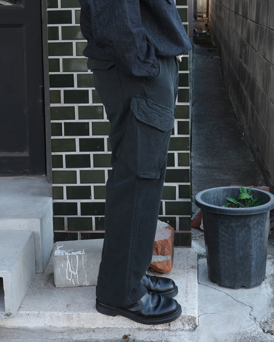 ミリタリートラウザーズをベースに、シルエットを現代的にリファイン。印象的な斜めのカーゴポケットが、立体感と実用性を両立させています。裾にかけて緩やかに絞られたテーパードラインにより、無骨ながらも洗練された印象に。素材には肌馴染みの良い10sヘリンボンを採用し、ストーンバイオウォッシュで柔らかな風合いとこなれ感を引き出しました。

Military trousers refined with a modern tapered silhouette. The distinctive slanted cargo pockets offer both practical utility and a structured look. Crafted from soft, classic 10s herringbone, this BLACK pair has been treated with a stone bio-wash, providing a comfortable, worn-in texture and a sophisticated take on rugged style.