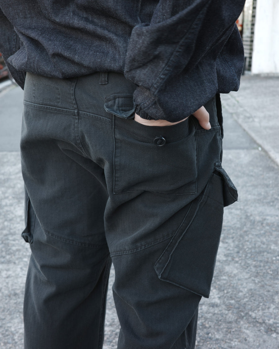 ミリタリートラウザーズをベースに、シルエットを現代的にリファイン。印象的な斜めのカーゴポケットが、立体感と実用性を両立させています。裾にかけて緩やかに絞られたテーパードラインにより、無骨ながらも洗練された印象に。素材には肌馴染みの良い10sヘリンボンを採用し、ストーンバイオウォッシュで柔らかな風合いとこなれ感を引き出しました。

Military trousers refined with a modern tapered silhouette. The distinctive slanted cargo pockets offer both practical utility and a structured look. Crafted from soft, classic 10s herringbone, this BLACK pair has been treated with a stone bio-wash, providing a comfortable, worn-in texture and a sophisticated take on rugged style.
