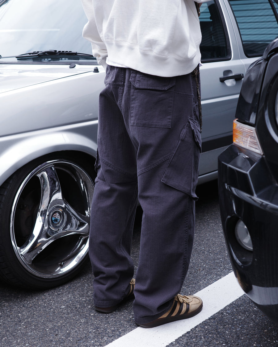 ミリタリートラウザーズをベースに、シルエットを現代的にリファイン。印象的な斜めのカーゴポケットが、立体感と実用性を両立させています。裾にかけて緩やかに絞られたテーパードラインにより、無骨ながらも洗練された印象に。素材には肌馴染みの良い10sヘリンボンを採用し、ストーンバイオウォッシュで柔らかな風合いとこなれ感を引き出しました。

Military trousers refined with a modern tapered silhouette. The distinctive slanted cargo pockets offer both practical utility and a structured look. Crafted from soft, classic 10s herringbone, this CHARCOAL pair has been treated with a stone bio-wash, providing a comfortable, worn-in texture and a sophisticated take on rugged style.