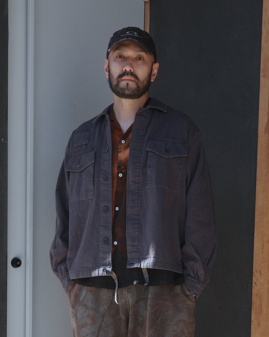 ミリタリーのシャツジャケットを現代的シルエットに更新。裾のドローコードで着丈のニュアンスを自在に調整可能です。素材には、やや甘織りで肌馴染みの良い10sヘリンボンを採用。ストーンバイオウォッシュを施すことで、クラシカルな生地感に柔らかな風合いとこなれ感をプラスした、着回し力の高い一着に仕上げました。

A modern update to the classic military shirt jacket, featuring hem drawcords for an adjustable silhouette. Crafted from 10s herringbone, slightly loose-knitted for a soft, comfortable touch. This BLACK piece has been treated with a stone bio-wash, giving the traditional weave a natural, worn-in texture and a sophisticated casual feel.